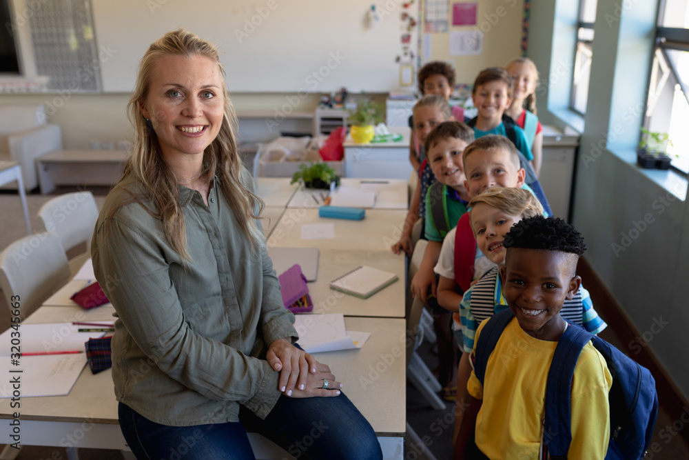 Female school teacher sitting on a desk in a classroom with a group of ...
