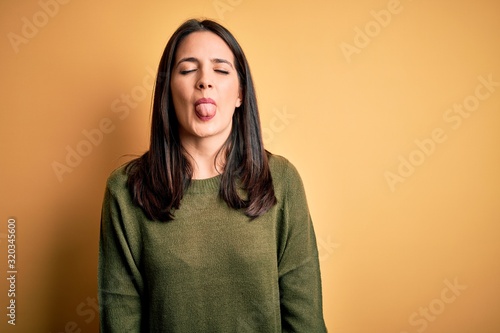 Obraz na plátně Young brunette woman with blue eyes wearing green casual sweater over yellow background sticking tongue out happy with funny expression