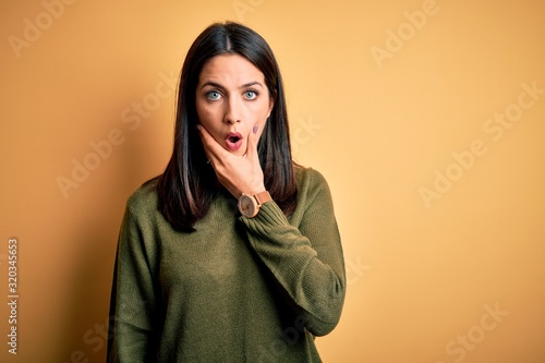 Young brunette woman with blue eyes wearing green casual sweater over yellow background Looking fascinated with disbelief, surprise and amazed expression with hands on chin © Krakenimages.com
