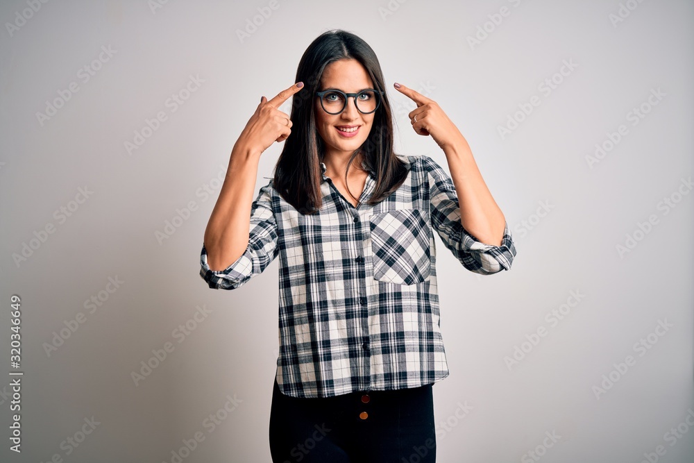 Young brunette woman with blue eyes wearing casual shirt and glasses over white background smiling pointing to head with both hands finger, great idea or thought, good memory Young brunette woman with blue eyes wearing casual shirt and glasses over white background smiling pointing to head with both hands finger, great idea or thought, good memory