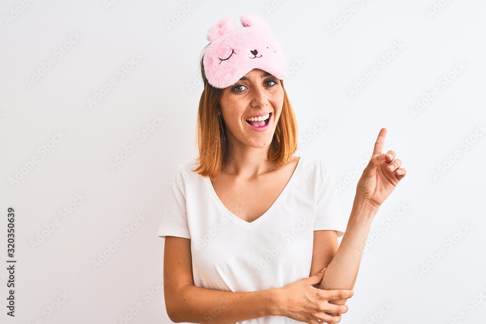 Beautiful redhead woman wearing sleeping mask over isolated background with a big smile on face, pointing with hand and finger to the side looking at the camera. Beautiful redhead woman wearing sleeping mask over isolated background with a big smile on face, pointing with hand and finger to the side looking at the camera.