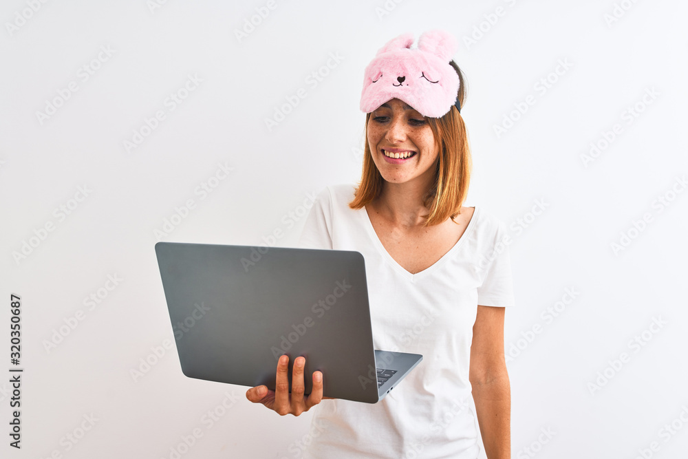 Beautiful redhead woman wearing sleeping mask using laptop over isolated background with a happy face standing and smiling with a confident smile showing teeth Beautiful redhead woman wearing sleeping mask using laptop over isolated background with a happy face standing and smiling with a confident smile showing teeth