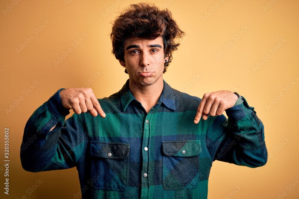 Young handsome man wearing casual shirt standing over isolated yellow background Pointing down looking sad and upset, indicating direction with fingers, unhappy and depressed. Young handsome man wearing casual shirt standing over isolated yellow background Pointing down looking sad and upset, indicating direction with fingers, unhappy and depressed.