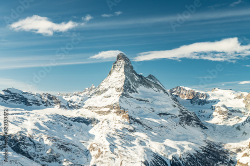 Scenic view on snowy Matterhorn (Cervin, Cervino) peak in sunny day with blue sky, pine trees, valley and clouds in background. Panoramic landscape in white and blue colors in Zermatt, Switzerland