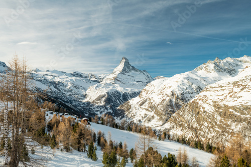 Scenic view on snowy Matterhorn (Cervin, Cervino) peak in sunny day with blue sky, pine trees, valley and clouds in background. Panoramic landscape in white and blue colors in Zermatt, Switzerland