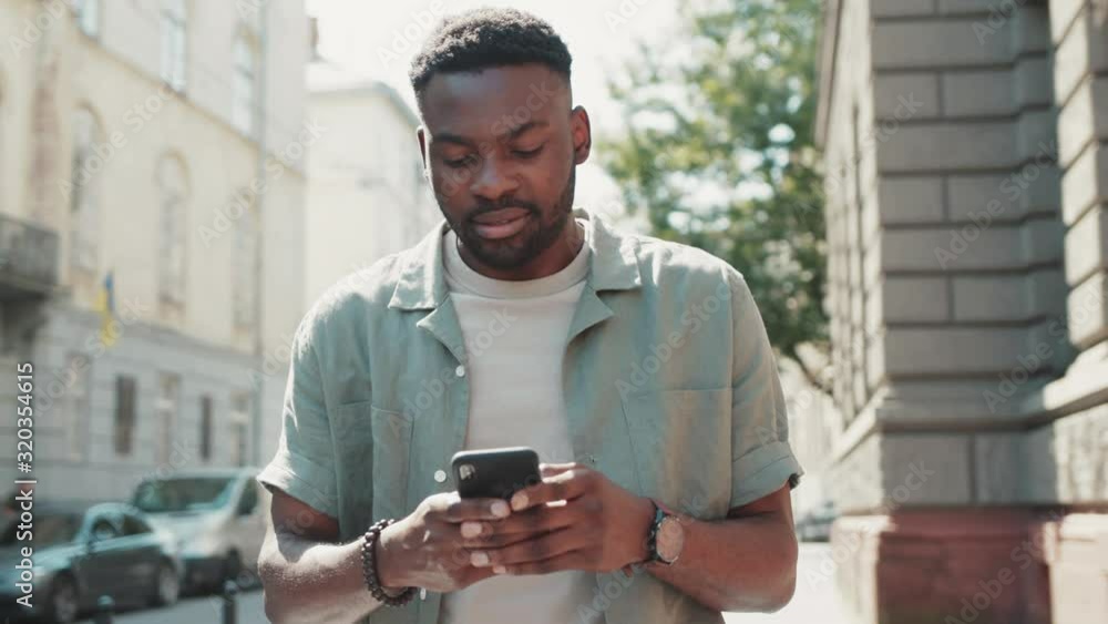 Happy attractive african american man walks down the street Typing on smartphone with interest Enjoying the walk Crowded city on the background sunset sunlight technology Happy attractive african american man walks down the street Typing on smartphone with interest Enjoying the walk Crowded city on the background sunset sunlight technology