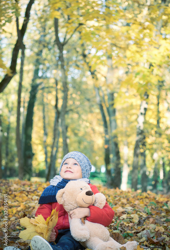 Little toddler boy  sitting with teddy bear in the autumn park  looking at throwing leaves around himself.
