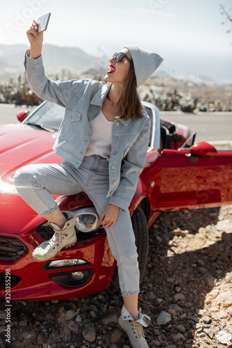 Young woman dressed casually with hat enjoying road trip on the island, standing with phone near the convertible car on the roadside