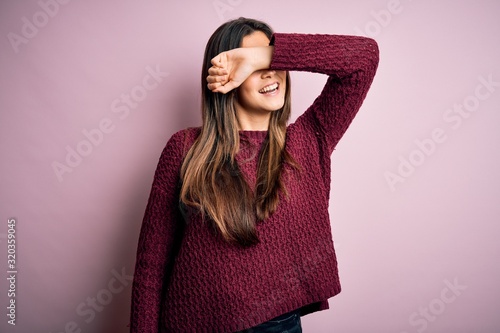 Young beautiful girl wearing casual sweater over isolated pink background covering eyes with arm smiling cheerful and funny. Blind concept.