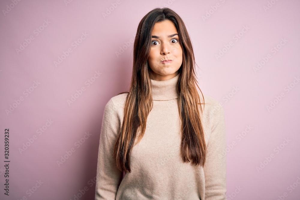 Young beautiful girl wearing casual turtleneck sweater standing over isolated pink background puffing cheeks with funny face. Mouth inflated with air, crazy expression. Young beautiful girl wearing casual turtleneck sweater standing over isolated pink background puffing cheeks with funny face. Mouth inflated with air, crazy expression.