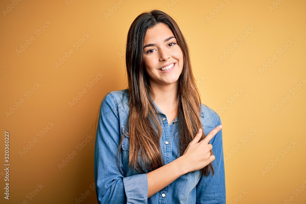 Young beautiful girl wearing casual denim shirt standing over isolated yellow background cheerful with a smile of face pointing with hand and finger up to the side with happy and natural expression Young beautiful girl wearing casual denim shirt standing over isolated yellow background cheerful with a smile of face pointing with hand and finger up to the side with happy and natural expression