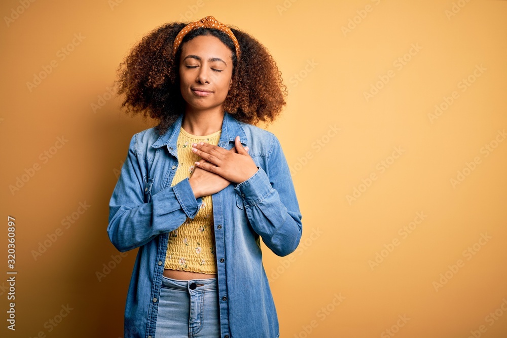 Beautiful african american with afro hair standing over yellow isolated background smiling with hands on chest with closed eyes and grateful gesture on face. Health concept. Beautiful african american with afro hair standing over yellow isolated background smiling with hands on chest with closed eyes and grateful gesture on face. Health concept.