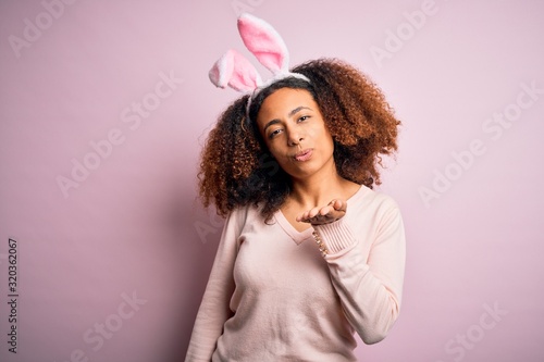 Young african american woman with afro hair wearing bunny ears over pink background looking at the camera blowing a kiss with hand on air being lovely and sexy. Love expression.