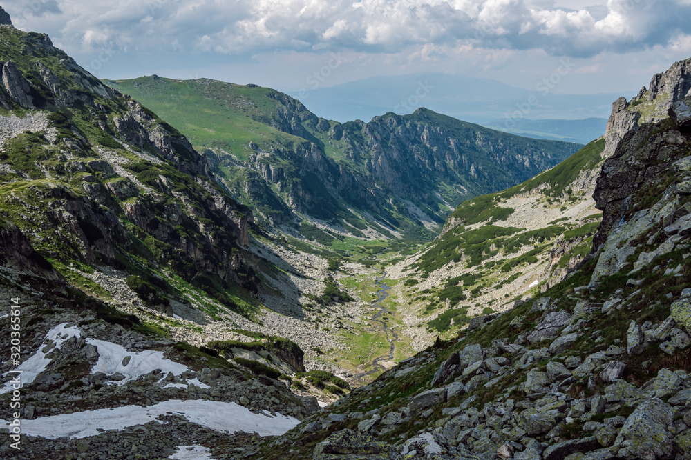 Panoramic view of a beautiful green valley with a flowing river through it and steep rocky slopes descending to it, in the background in the distance is Vitosha Mountain, Rila Mountain, Bulgaria