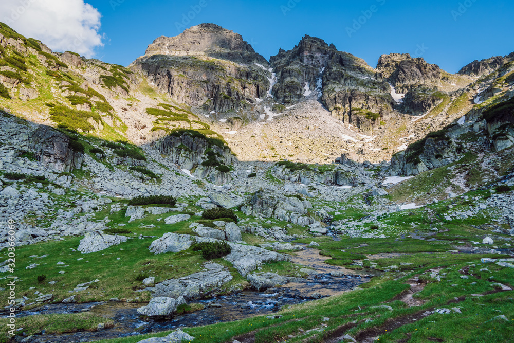 View of a beautiful green mountain valley with small river running through it and high rocky peaks hanging over it, Malyovitsa hiking trail, Rila mountain, Bulgaria.