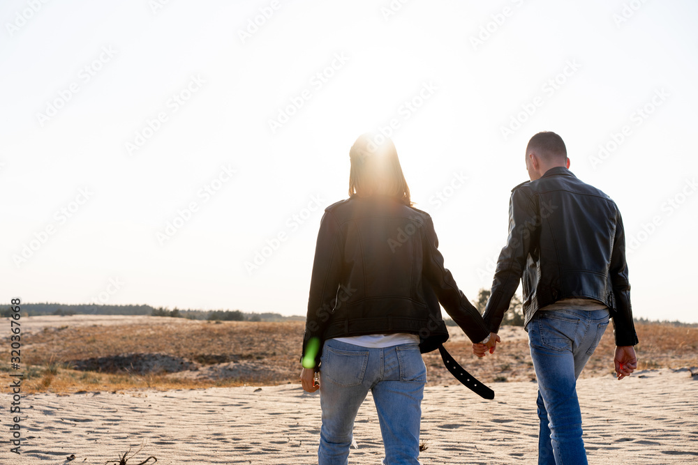 Young European couple dressed leather jacket and blue jeans walking desert holding hands view from the back. Young European couple dressed leather jacket and blue jeans walking desert holding hands view from the back.