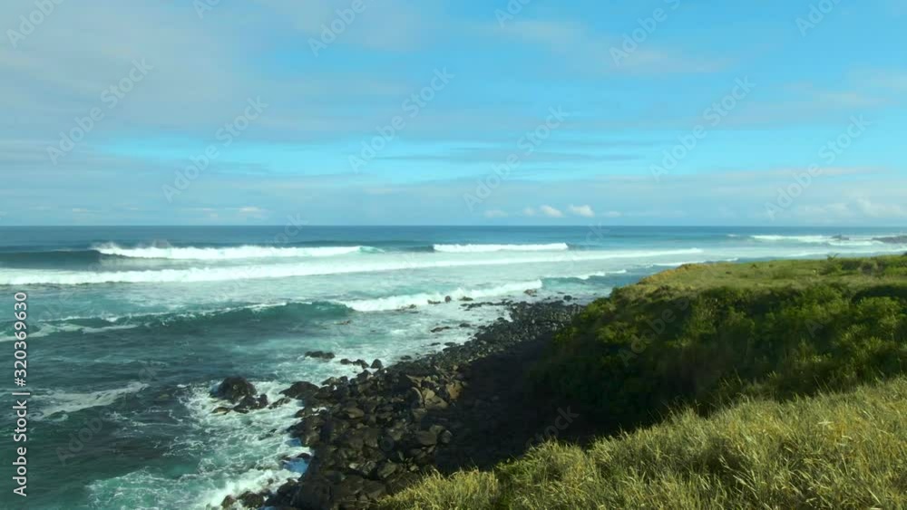 Green grass covering coastline of Hawaiian island, big white waves splashing over rocky shore on a sunny day