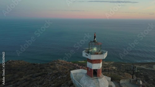 Young male carefully standing on top of a lighthouse on a mountain looking out over the ocean and Cabo San Lucas, Mexico, at sunset. Aerial parallax. photo