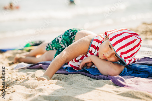 Summer portrait of cute little boy relaxing on sand beach by the sea, lying down covered with towels, vacation with children