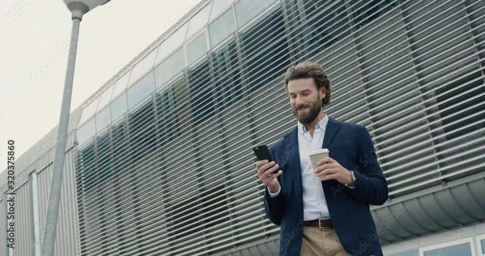 Young bearded Caucasian Man is standing near Industrial Building with Phone. Chatting on his Smartphone and drinking Coffee to go. Stylish Businessman with Phone. Apps. Social Network. People. Young bearded Caucasian Man is standing near Industrial Building with Phone. Chatting on his Smartphone and drinking Coffee to go. Stylish Businessman with Phone. Apps. Social Network. People.