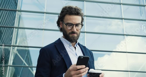 Young confident Professor in Glasses looking to the Camera confidently. Portrait of young Caucasian Man with stylish hair and Look. Using his Smartphone. Social Network. Work outside. Success. People.