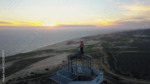 Young male standing on top of a lighthouse looking out over Cabo San Lucas, Mexico, at sunset. Aerial push in photo