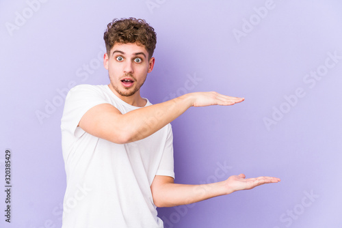 Young blond curly hair caucasian man isolated shocked and amazed holding a copy space between hands.