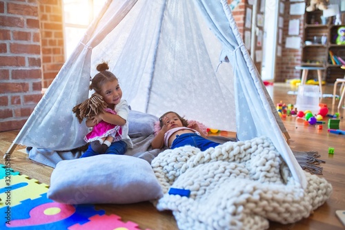 Adorable toddlers lying down over blanket inside tipi smiling and playing with doll at kindergarten