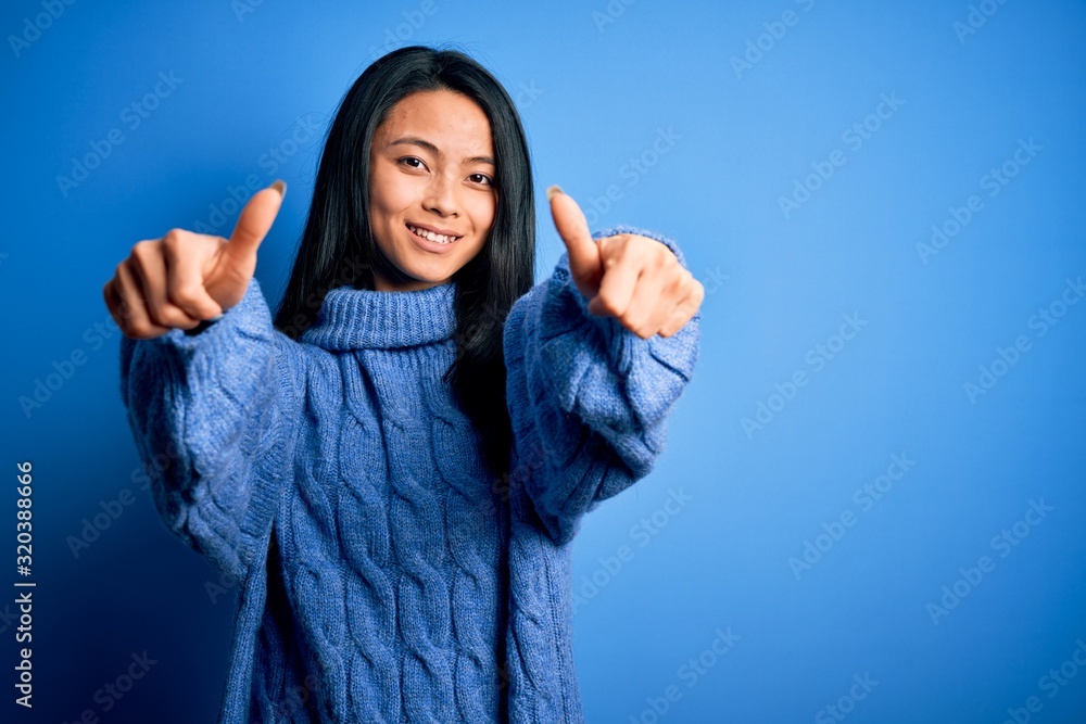 Young beautiful chinese woman wearing casual sweater over isolated blue background approving doing positive gesture with hand, thumbs up smiling and happy for success. Winner gesture. Young beautiful chinese woman wearing casual sweater over isolated blue background approving doing positive gesture with hand, thumbs up smiling and happy for success. Winner gesture.