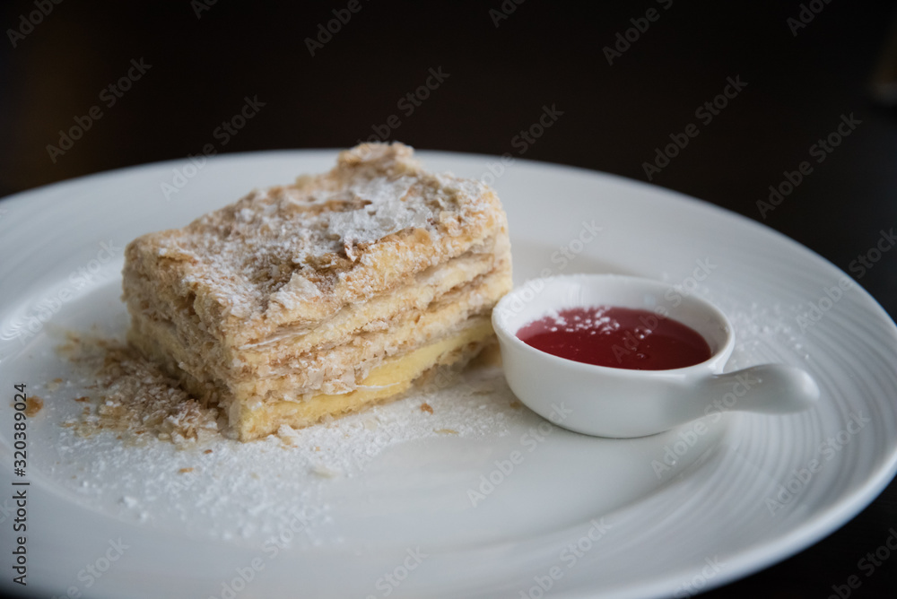 A piece of Napoleon cake on a white plate. The cake is sprinkled with powdered sugar. There is a Cup of jam next to it. A piece of Napoleon cake on a white plate. The cake is sprinkled with powdered sugar. There is a Cup of jam next to it.