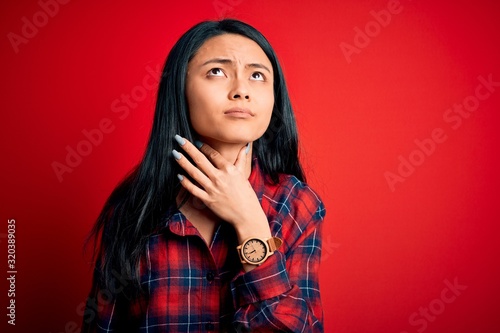 Young beautiful chinese woman wearing casual shirt over isolated red background Touching painful neck, sore throat for flu, clod and infection © Krakenimages.com