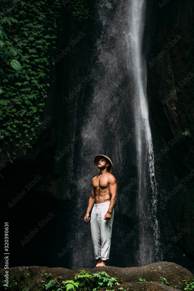 A man of athletic build at the waterfall. A man travels the world. Man at the waterfall. Travel to Bali Indonesia. A man in a conical hat and white pants at the waterfall. Lone traveler. A man of athletic build at the waterfall. A man travels the world. Man at the waterfall. Travel to Bali Indonesia. A man in a conical hat and white pants at the waterfall. Lone traveler.
