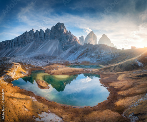 Aerial view of beautiful rocks  mountain lake  reflection in water and houses on the hill at sunset. Autumn landscape with mountains  blue sky and sunlight. Dolomites  Italy. Top view of Italian alps