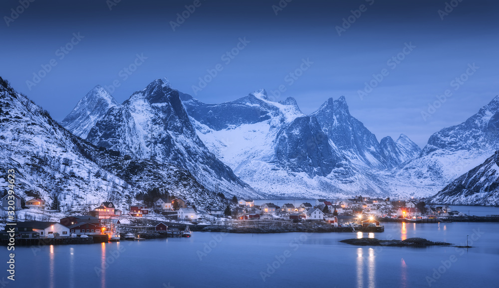 Houses in village, city lights, snowy mountains, sea, blue cloudy sky reflected in water at night in winter. Landscape with beautiful Reine at night in Lofoten islands, Norway. Norwegian rorbu at dusk