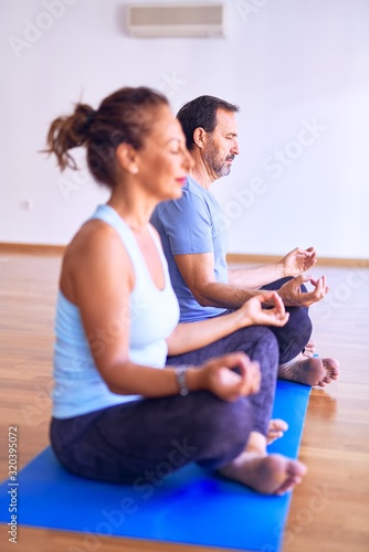 Middle age beautiful sporty couple sitting on mat practicing yoga doing lotus pose at gym