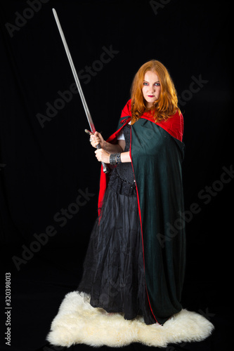 red-haired girl wearing in a long black gown and green robe with two-handed sword standing on a white sheepskin photo