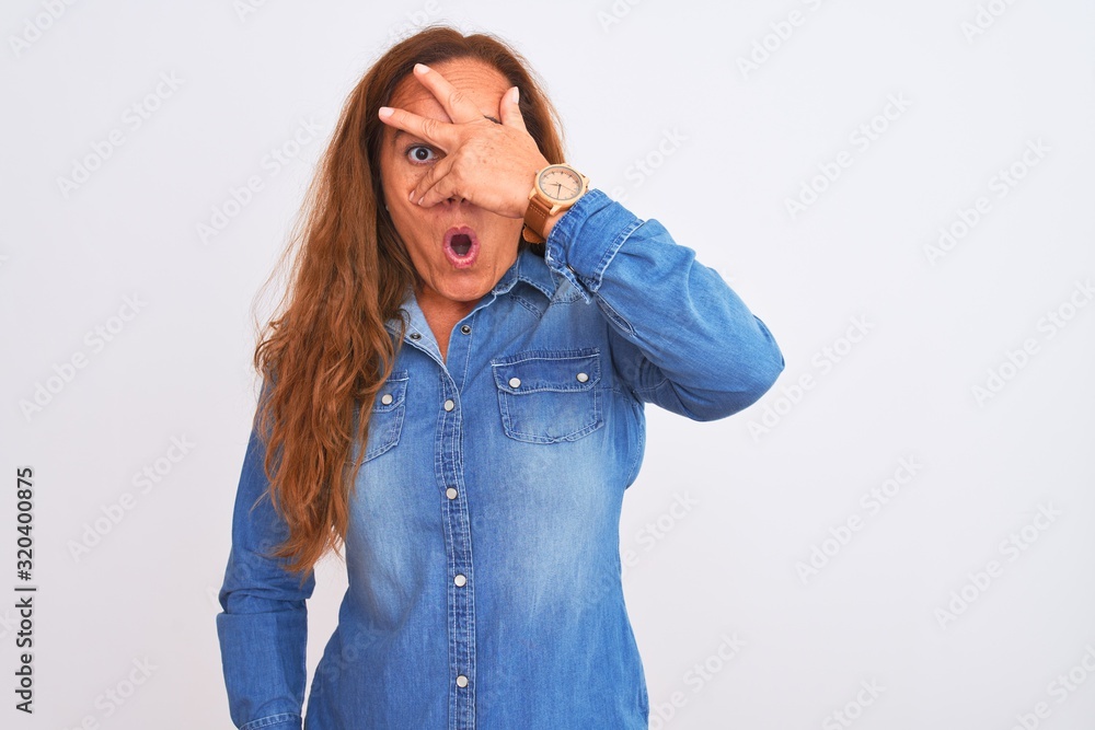 Middle age mature woman wearing denim jacket standing over white isolated background peeking in shock covering face and eyes with hand, looking through fingers with embarrassed expression. Middle age mature woman wearing denim jacket standing over white isolated background peeking in shock covering face and eyes with hand, looking through fingers with embarrassed expression.