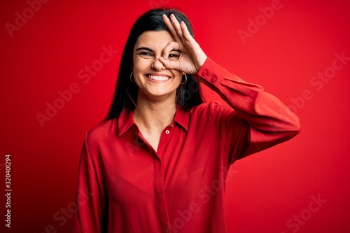 Young beautiful brunette woman wearing casual shirt standing over red background doing ok gesture with hand smiling, eye looking through fingers with happy face. © Krakenimages.com
