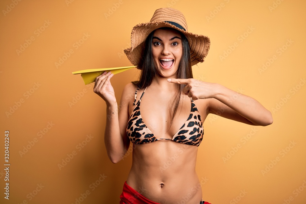 Young beautiful woman on vacation wearing bikini and summer hat holding paper airplane very happy pointing with hand and finger Young beautiful woman on vacation wearing bikini and summer hat holding paper airplane very happy pointing with hand and finger