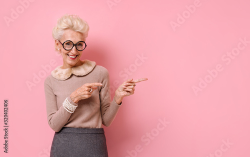 Smiling woman points away with both forefingers, shows on copy space for your text. Photo of kind elderly woman on pink background. photo