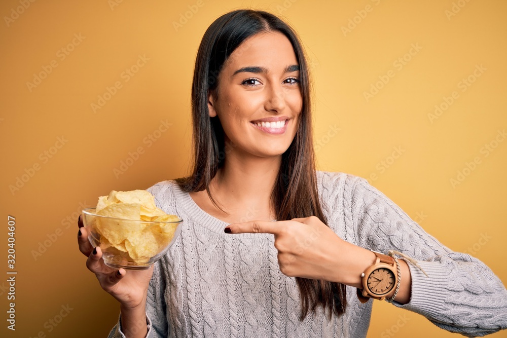 Young beautiful brunette woman holding bowl with snack potato chips over yellow background very happy pointing with hand and finger Young beautiful brunette woman holding bowl with snack potato chips over yellow background very happy pointing with hand and finger