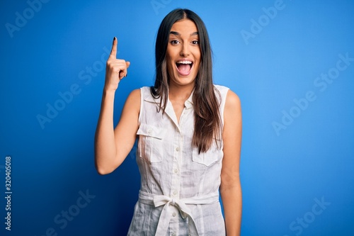 Young beautiful brunette woman wearing casual dress over isolated blue background pointing finger up with successful idea. Exited and happy. Number one.