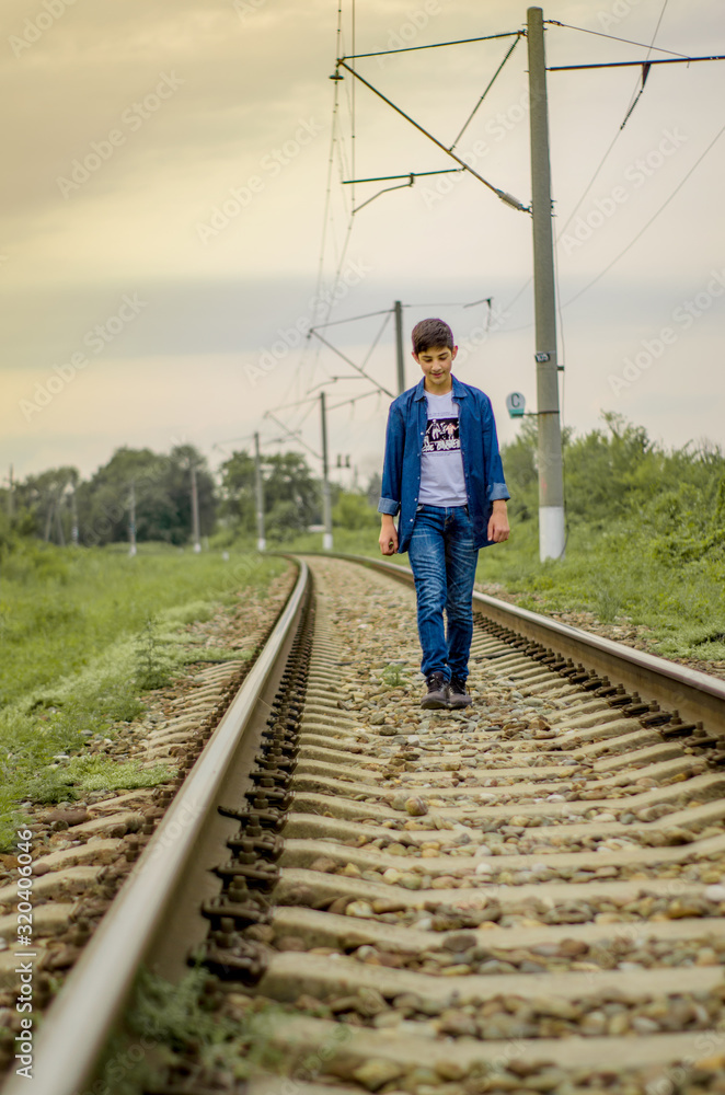 A dark-haired guy in a denim shirt and jeans walks along a railroad track on sleepers at sunset. The guy looks at the camera. Selective focus.
