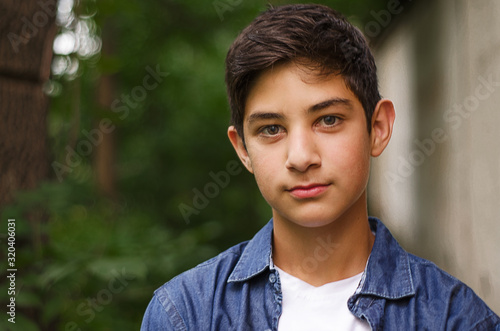 Portrait of a dark-haired teenager in a denim shirt against a background of greenery. The guy looks at the camera. Selective focus. Close up.