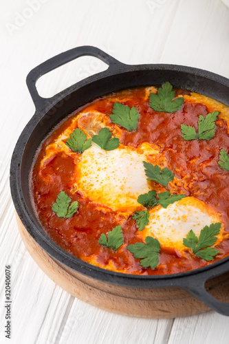 Shakshuka - a dish of eggs fried in a sauce of tomato, hot pepper, onion and seasoning. Shakshuka Scrambled eggs with tomatoes and vegetables on white wooden background. Breakfast in a Frying Pan.