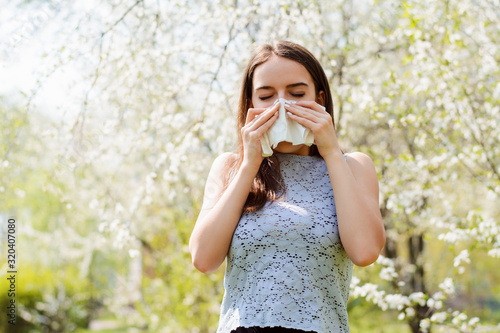 Sneezing young girl with handkerchief among blooming trees in spring park. Concept of allergy