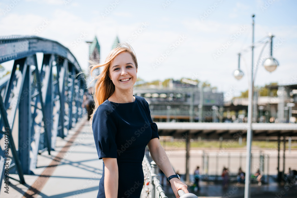 Portrait of a young happy woman standing at bridge near station. Attractive blonde arrived to some new city expresses happiness and joy of traveling