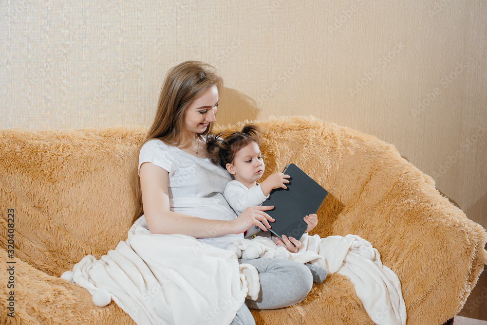 A pregnant mother is sitting on the couch with her little daughter and playing with a gadget A pregnant mother is sitting on the couch with her little daughter and playing with a gadget