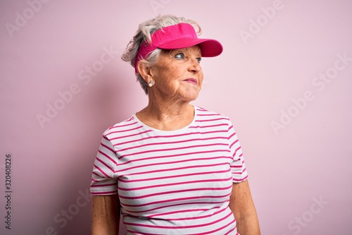 Senior beautiful sporty woman wearing sport cap standing over isolated pink background looking away to side with smile on face, natural expression. Laughing confident.