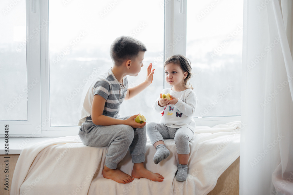 Brother and sister are sitting on the windowsill playing and eating apples. Happiness Brother and sister are sitting on the windowsill playing and eating apples. Happiness
